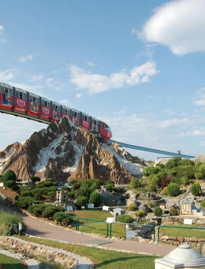 Treno sospeso sopra un parco con miniature di montagne e edifici.